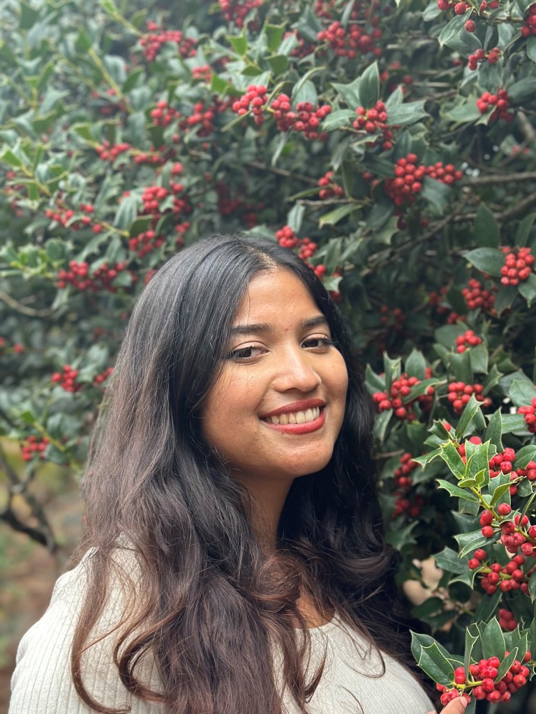 A smiling woman with long, wavy hair stands next to a holly bush, adorned with bright red berries.