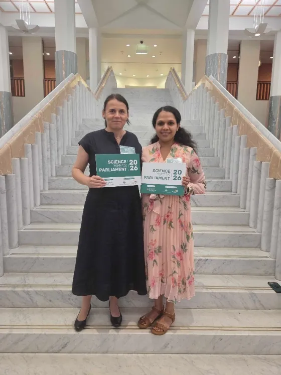 Two women stand on a staircase, holding up signs that read 'Science in Parliament 2026.' One woman wears a black dress, and the other is dressed in a floral pink outfit.
